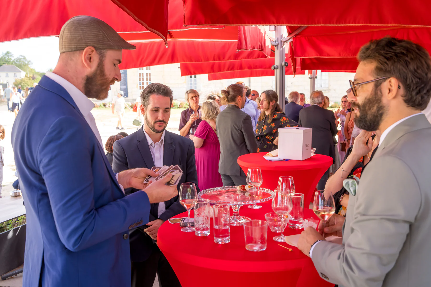 Ugo avec un H montre un jeu de cartes à des invités autour d'une table haute, sous des parasols rouges