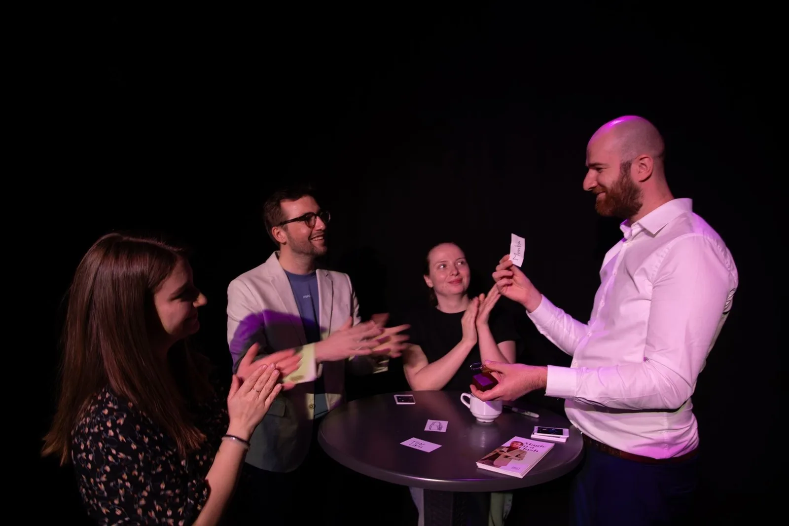 Trois spectateurs applaudissent autour d'une table haute pendant qu'Hugo montre une carte